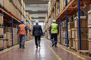 Three workers walking down the aisle of a warehouse, with shelves stacked with boxes on either side of the aisle