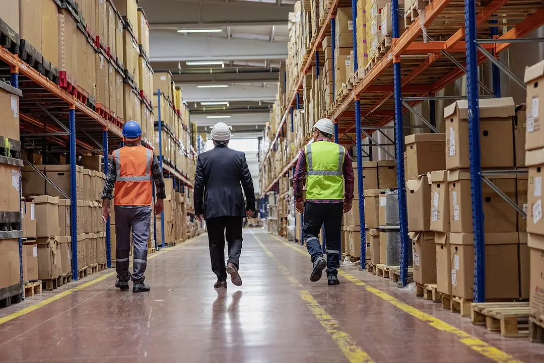 Three workers walking down the aisle of a warehouse, with shelves stacked with boxes on either side of the aisle