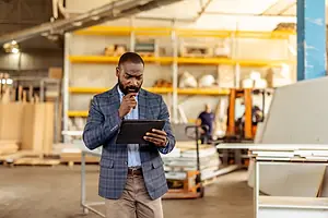 A man in a business suit standing in a warehouse, while holding an iPad and looking at it thoughtfully