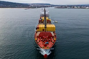 A red container ship sails through calm waters, low hills in the background