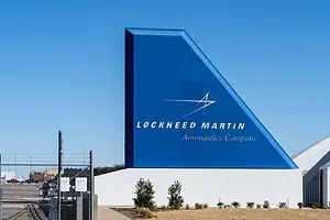 A large blue sign shaped by an airplane tailfin outside of a gated facility, the reads "LOCKHEED MARTIN"
