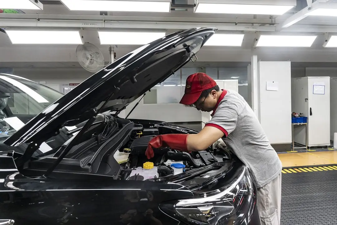 AN AUTO WORKER LEANS UNDER THE HOOD OF A CAR, WEARING RED RUBBER GLOVES